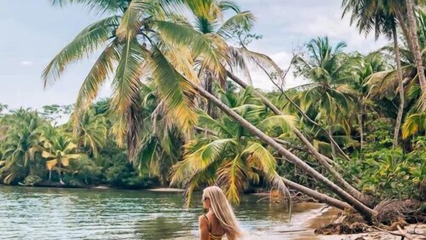 A woman in a yellow dress standing on a sandy beach with palm trees and a river in the background.