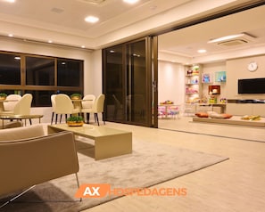A modern living room with a sofa, chairs, a coffee table, and a TV. There is a bookshelf with toys and a clock on the wall.