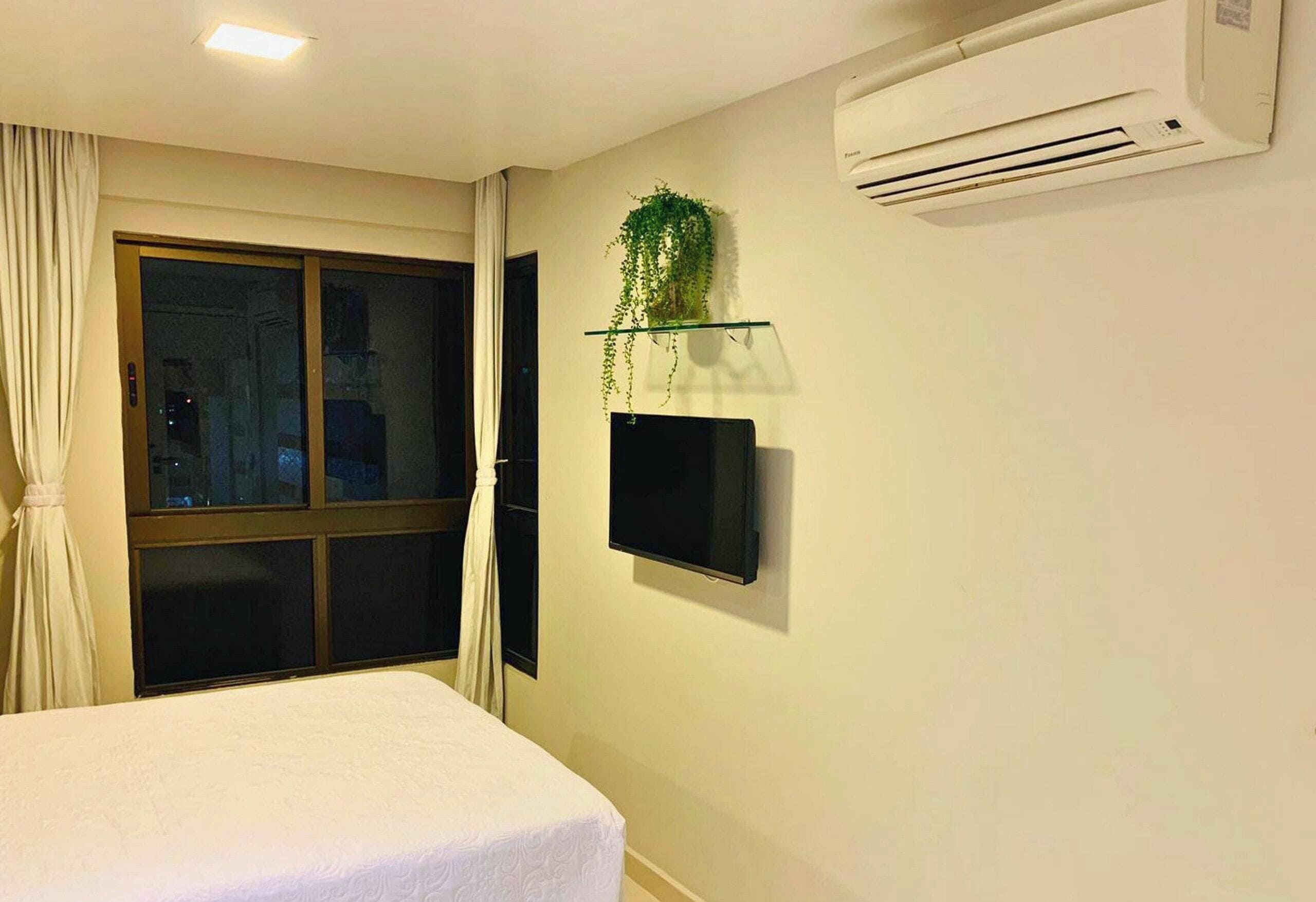 A hotel room with a bed, a TV, an air conditioner, a window with curtains, and a potted plant on a shelf.