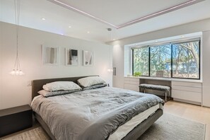 Un dormitorio moderno con una cama grande, un banco y una ventana con vistas a árboles.