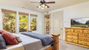 A bedroom with a bed, a wooden dresser, a ceiling fan, and large windows with a view of trees.
