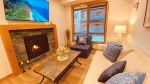 A living room with a stone fireplace, a flat-screen TV, a sofa with patterned cushions, and a window with blinds. - Luxury, Spacious, New, Entire Condo Suite, Scenery Year-round Mountain Getaway (Canmore)