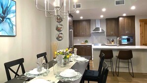 A dining area with a long table set for six, a chandelier, and a kitchen in the background. - Luxury, Spacious, New, Entire Condo Suite, Scenery Year-round Mountain Getaway (Canmore)