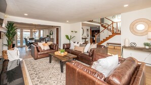 Ein großzügiger Wohnraum mit Ledersofas, einem Couchtisch und einem gemusterten Teppich. Eine Treppe führt zu einer oberen Ebene.
