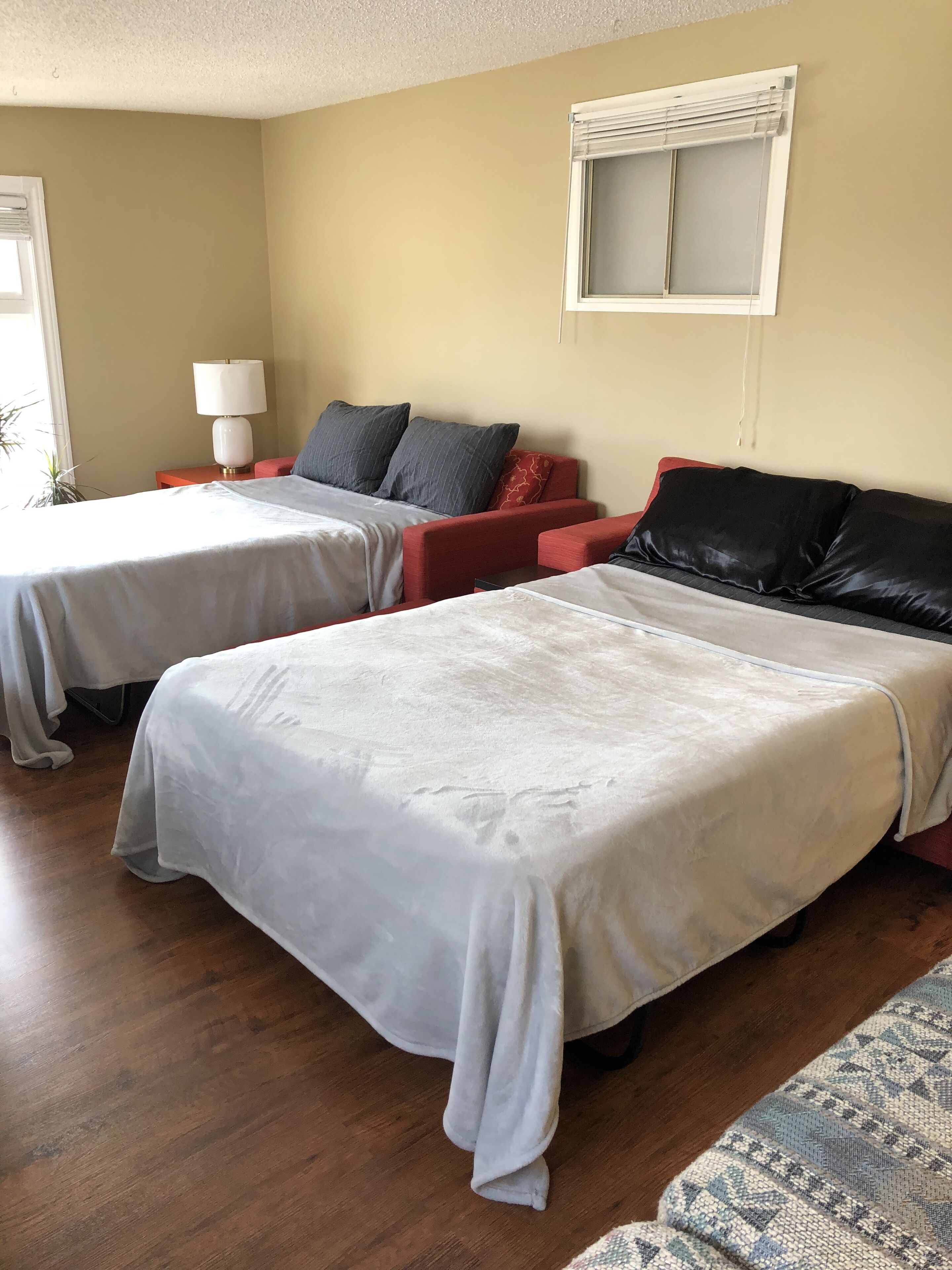 A hotel room with two beds, a red sofa, a lamp, and a window with blinds.
