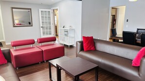 Ein modernes Wohnzimmer mit einem braunen Ledersofa, einer rot gepolsterten Bank und einem kleinen Holztisch. Außerdem ist ein weißes Bücherregal und ein Spiegel zu sehen.