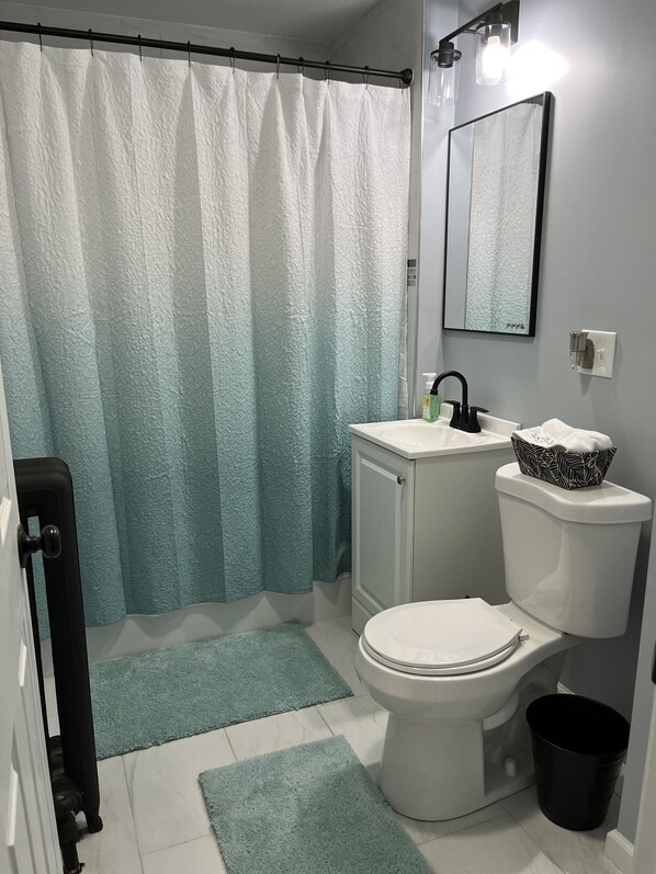 A bathroom with a white toilet, a sink with a black faucet, a mirror above the sink, a shower curtain, and a black trash bin.
