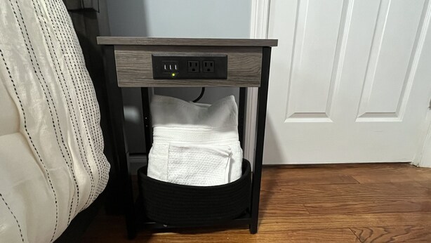 A bedside table with a power strip, a white towel, and a black basket. - Upscaled Quiet Neighborhood- Modern 3 BDRM (Springfield)