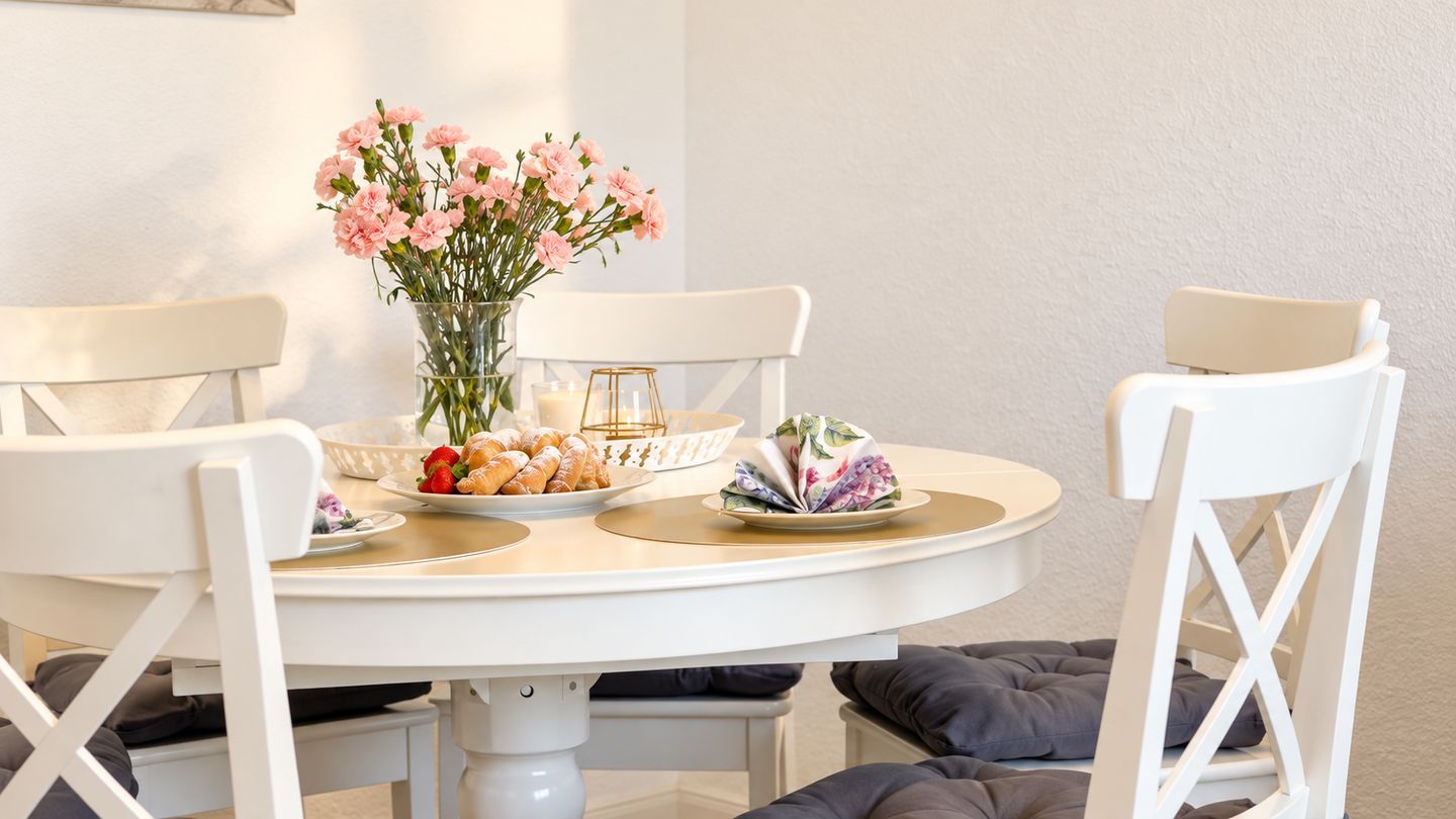 Ein runder Esstisch mit weißen Stühlen, eine Vase mit rosafarbenen Blumen und angerichteten Speisen.