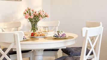 Ein runder Esstisch mit weißen Stühlen, eine Vase mit rosafarbenen Blumen und angerichteten Speisen.