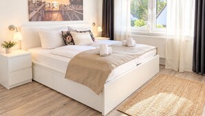 Un dormitorio moderno con una cama grande, una mesita de noche con una lámpara, una ventana con cortinas y un cuadro en la pared.