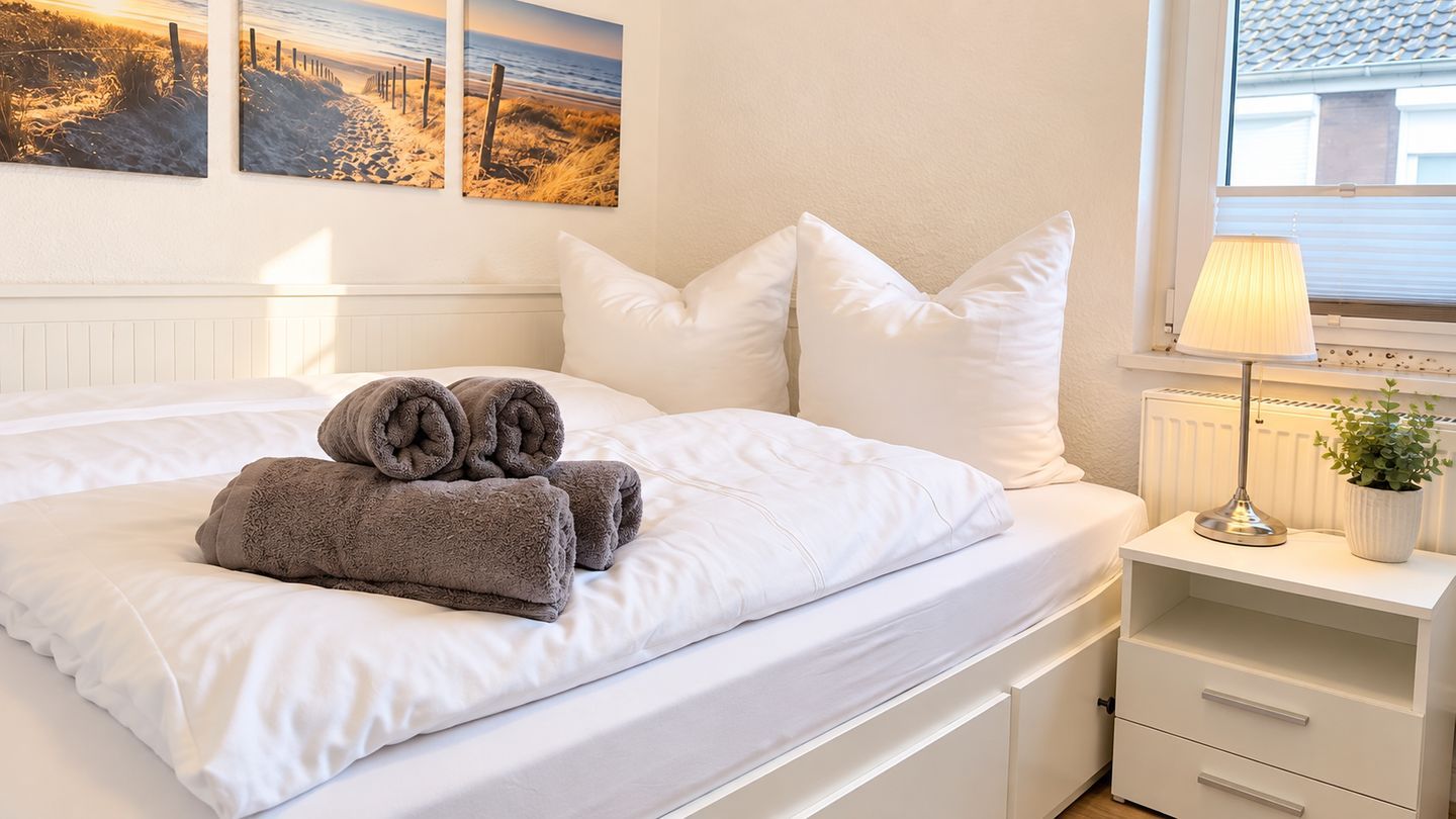 Ein ordentlich bezogenes Bett mit weißer Bettwäsche und einem Stapel gefalteter Handtücher, ein Nachttisch mit einer Lampe und einer Topfpflanze sowie ein Fenster mit Blick auf den Strand.