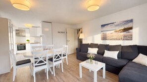 Ein modernes Wohnzimmer mit einem dunkelgrauen Sofa, einem weißen Esstisch und Stühlen. An der Wand hängt ein Gemälde und auf einem kleinen Tisch steht eine Vase mit Blumen.