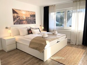 Un dormitorio con cama, mesitas de noche, una ventana con cortinas y un cuadro en la pared.