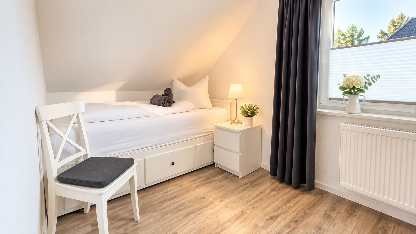 Ein Schlafzimmer mit Bett, Nachttisch, Lampe, Stuhl und einem Fenster mit Vorhängen.