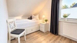 Un dormitorio con cama, mesita de noche, lámpara, silla y ventana con cortinas.