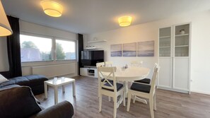 Una sala moderna con un sofá, una mesa de centro, una mesa de comedor con sillas, un televisor y una zona de cocina con un refrigerador.