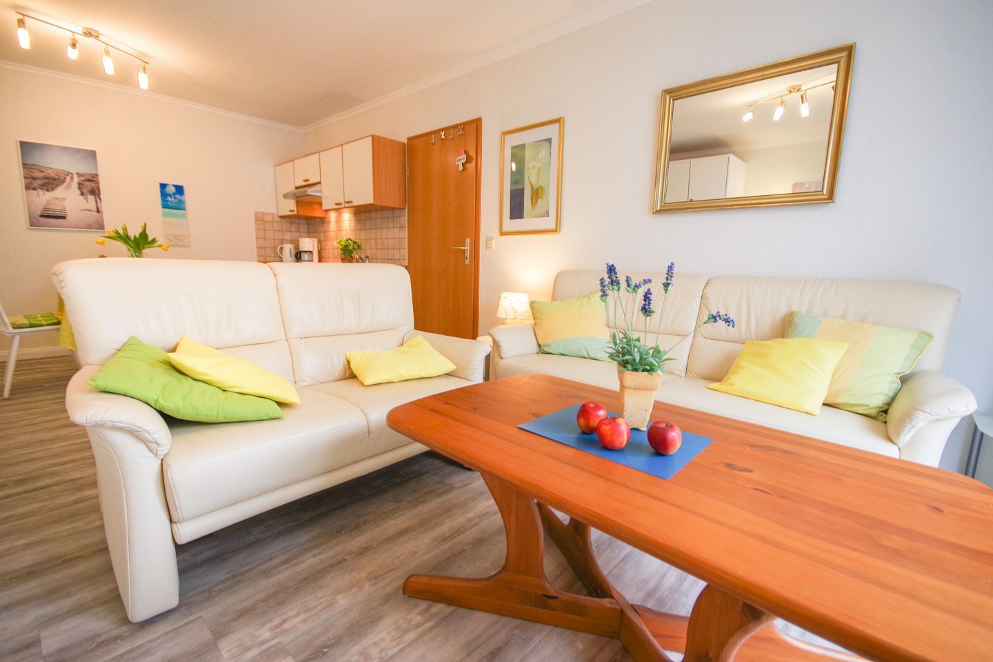 A living room with a wooden coffee table, two white sofas with cushions, a wooden dining table, and a kitchen area in the background.