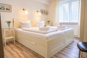 A double bed with white linens and towels, bedside tables with lamps, a window with curtains, and a wooden floor.