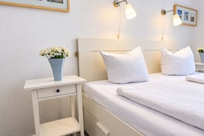 Un lit bien fait, avec du linge de lit et des oreillers blancs, une table de chevet ornée d’un vase de fleurs et des lampes fixées au mur.