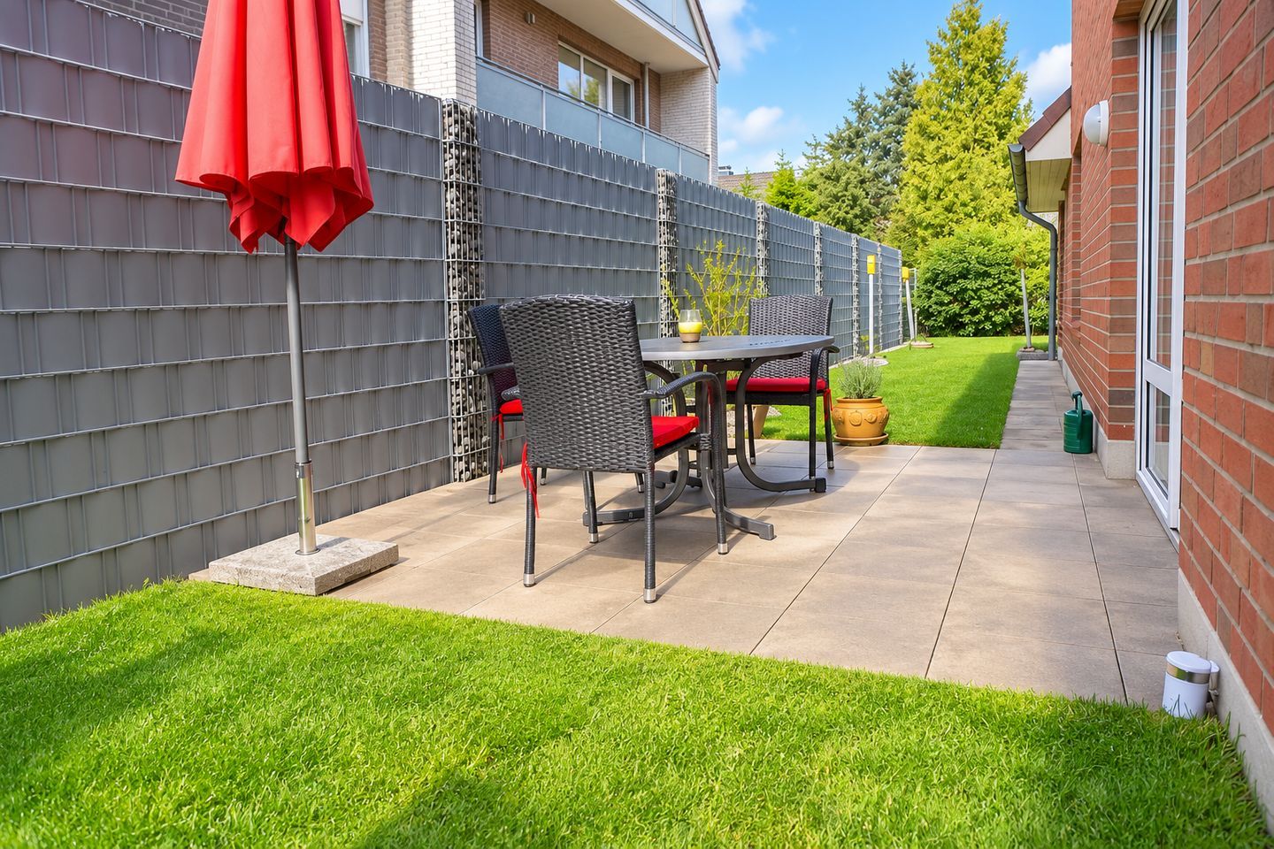Eine Terrasse mit Tisch, Stühlen und einem roten Sonnenschirm, an einer Ziegelwand.