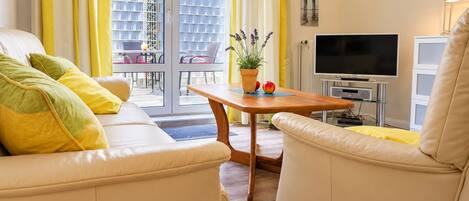 A modern living room with a sofa, armchairs, a coffee table, a TV, and a balcony with yellow curtains.