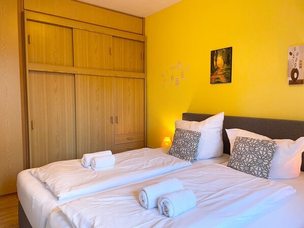 A double bed with white linens and rolled towels, a wooden wardrobe, and a yellow wall with a painting and wall art.