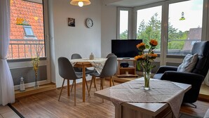 A modern living room with a dining table, chairs, a TV, and a large window with a view of a building and trees.