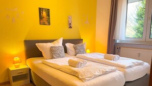 A double bed with white linens, two pillows, a bedside table with a lamp, and a window with curtains.