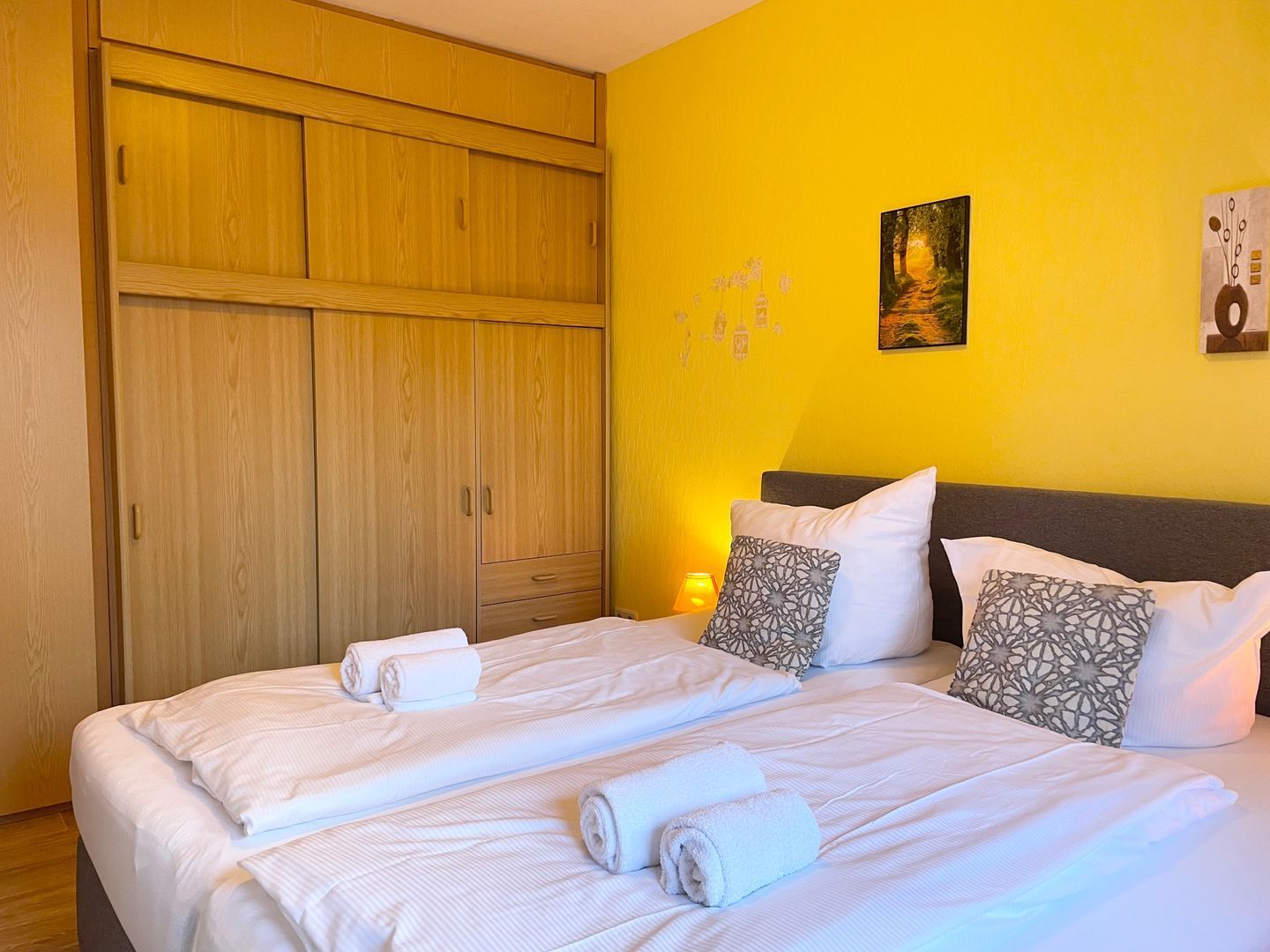 Un lit double avec du linge de lit blanc et des serviettes enroulées, une armoire en bois et un mur jaune orné d’un tableau et d’autres éléments décoratifs.