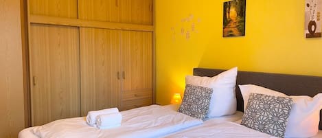 Cama doble con sábanas blancas y toallas enrolladas, armario de madera y pared amarilla con un cuadro y decoración mural.