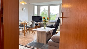 A room with a dining table, chairs, a TV, a sofa, and a window with a view of greenery.