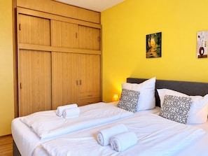 Una cama doble con sábanas blancas y toallas enrolladas, un armario de madera y una mesita de noche con lámpara.