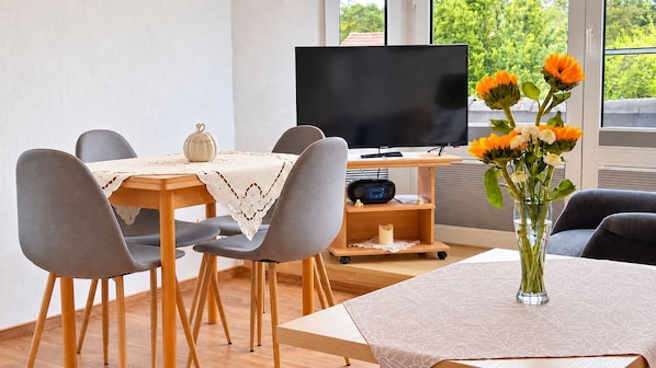 A modern living room with a dining area, a flat-screen TV, and a vase of flowers.