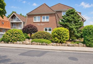 Un edificio residencial de dos pisos con fachada de ladrillo, múltiples ventanas y un jardín bien cuidado con arbustos variados y un gran árbol de coníferas.