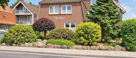 A two-story residential building with a brick facade, multiple windows, and a well-maintained garden with various shrubs and a large coniferous tree.