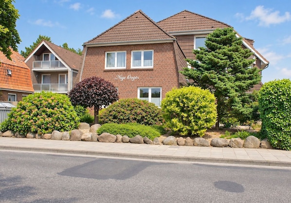 A two-story residential building with a brick facade, multiple windows, and a well-maintained garden with various shrubs and a large coniferous tree.