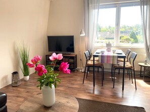 Una sala de estar con televisor, mesa de comedor, sillas, mesa de centro, planta en maceta y ventana con cortinas.