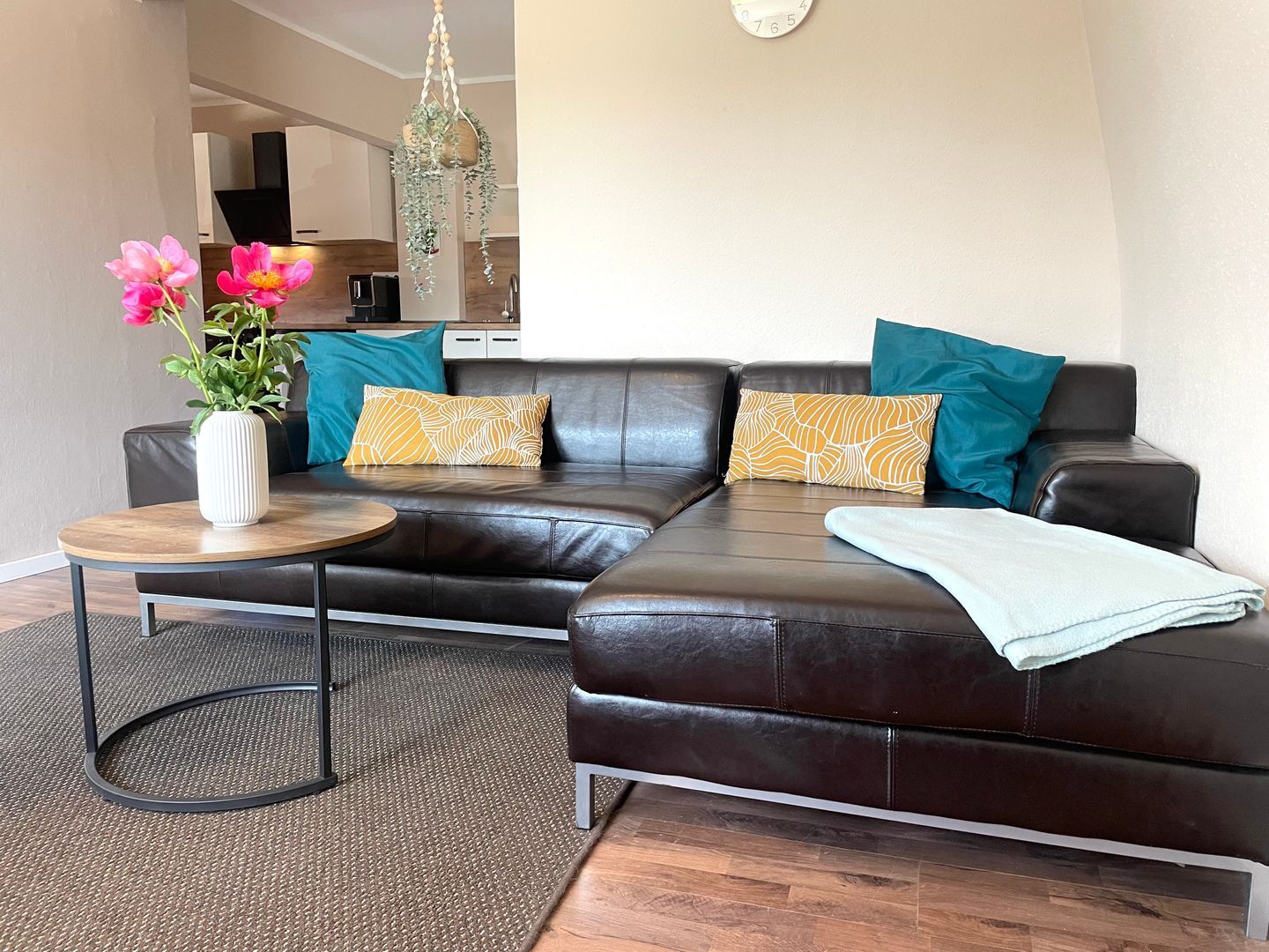 A modern living room with a leather sofa, a round wooden coffee table, a vase with flowers, and a hanging plant.
