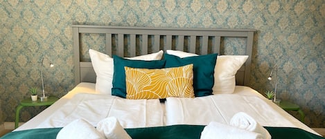 Una cama bien hecha con sábanas blancas, almohadas verdes y amarillas, y dos toallas enrolladas sobre una colcha verde.