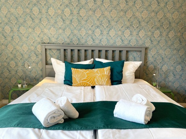A neatly made bed with white linens, green and yellow pillows, and two rolled towels on a green bedspread.