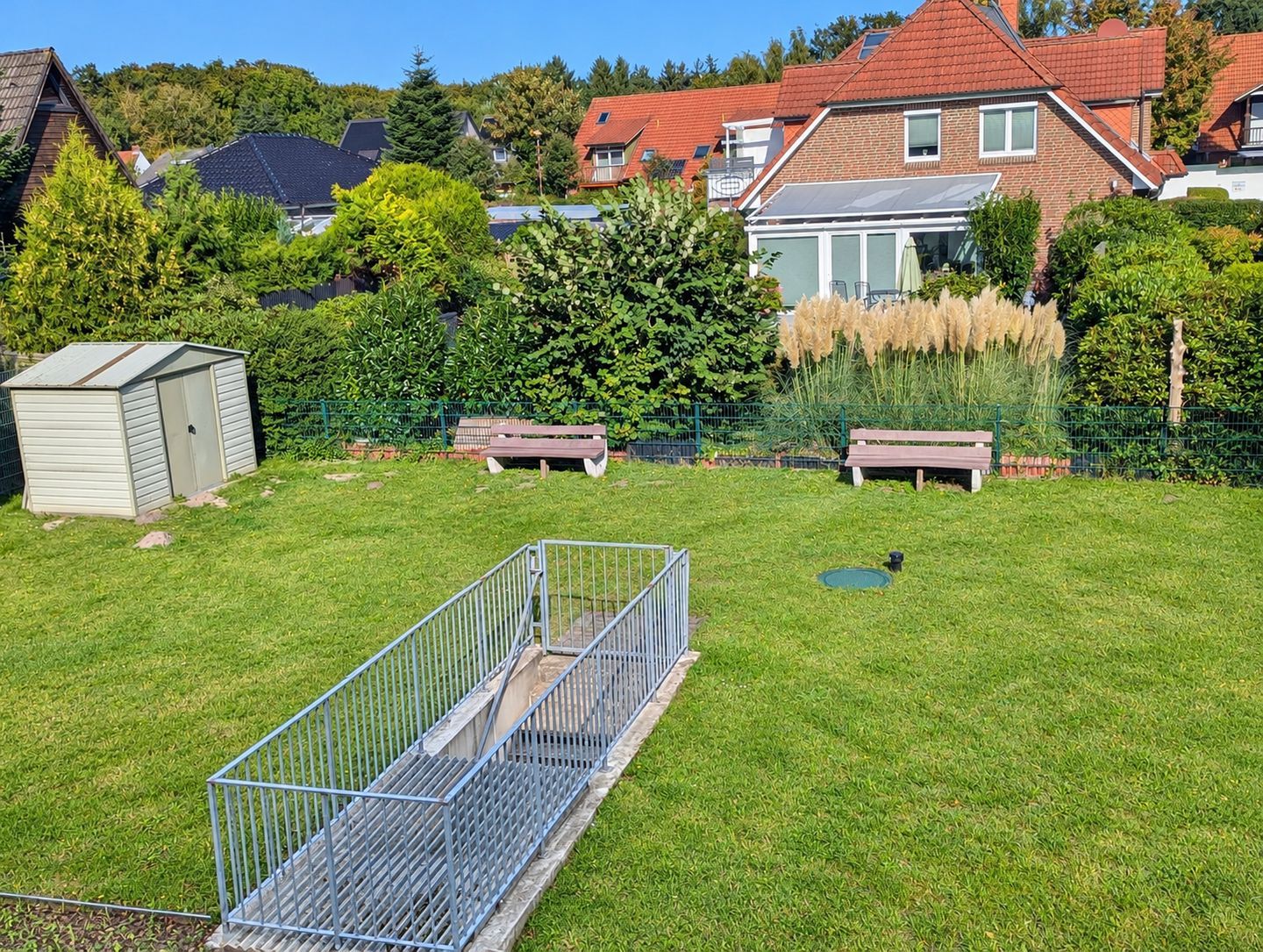 Ein Wohnhausgarten mit Metallzaun, einer kleinen Gartenhütte und zwei Bänken.