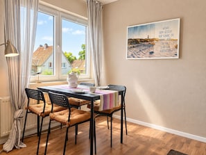 A dining area with a table set for four, chairs, a window with curtains, and a wall-mounted picture. - NO6 - Ferienwohnung mit Gemütlicher Ausstattung, 5 Min. zum Strand - Nige Ooge (Cuxhaven)