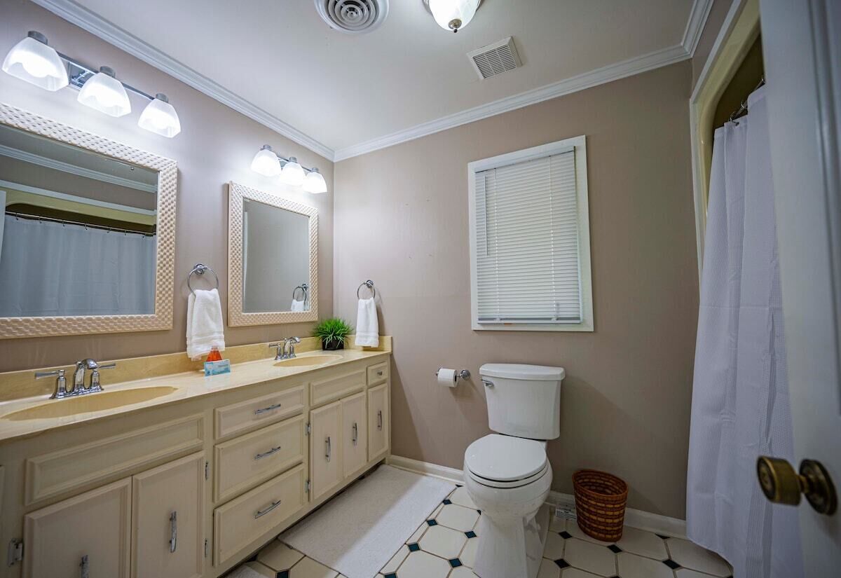 A bathroom with a double sink vanity, a toilet, a mirror, and a window with blinds.