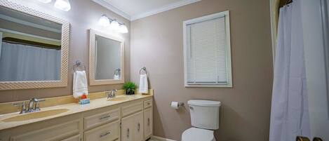 A bathroom with a double sink vanity, a toilet, a mirror, and a window with blinds.
