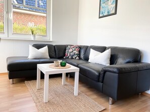 Um sofá modular de couro preto com almofadas brancas, uma pequena mesa de centro branca com objetos decorativos, um tapete tecido, uma janela com vista para um edifício de tijolos e um quadro na parede.