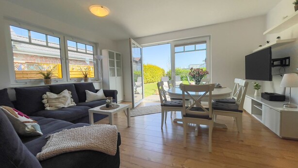 A spacious living room with a dark blue sofa, a coffee table, and a dining area with a white table and chairs. There is a kitchen area with a white countertop and a TV mounted on the wall.