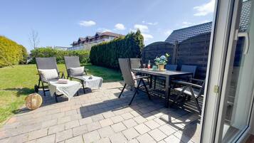 Een terras buiten met rieten stoelen, een tafel en een klein rond tafeltje met een fles en bloemen.