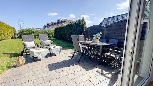 An outdoor patio with wicker chairs, a table, and a small round table with a bottle and flowers.