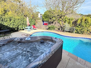 Un jacuzzi junto a una piscina con zona de patio, rodeado de árboles y una valla de madera.